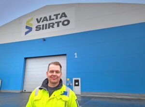 Kuvassa iso varastohalli, jonka edustalla mies kirkkaan neonkeltaisessa turvatakissa