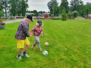 Maisema Harjavallan Viuhka-aukion viereiseltä nurmialueelta. Kuvassa kaksi lippispäistä lasta pelaa jalkapalloa.