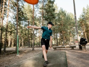 Mies heittää punaista frisbee-kiekkoa tummansinisessä shortsiasussa. Taustalla mäntymetsää ja maa mäntykangasta.