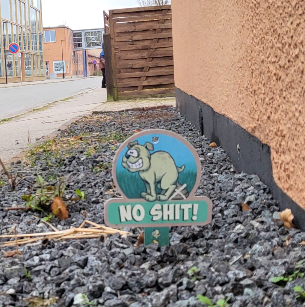Kuva talon vierestä, maasta jossa on soraa, johon tökätty kyltti. Pyöreänmuotoisessa kyltissä kuva koirasta vihreällä nurmikolla sinisen taivaan alla ja lukee "No shit!"